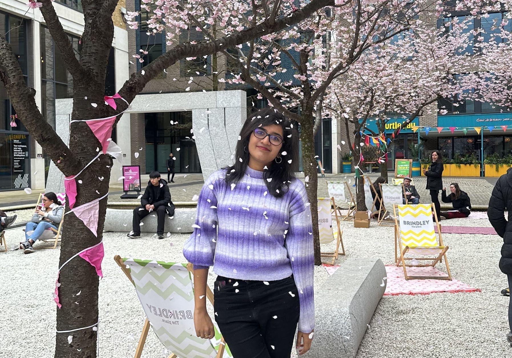 A female student wearing a purple striped jumper and black jeans stands in front of a cherry blossom tree