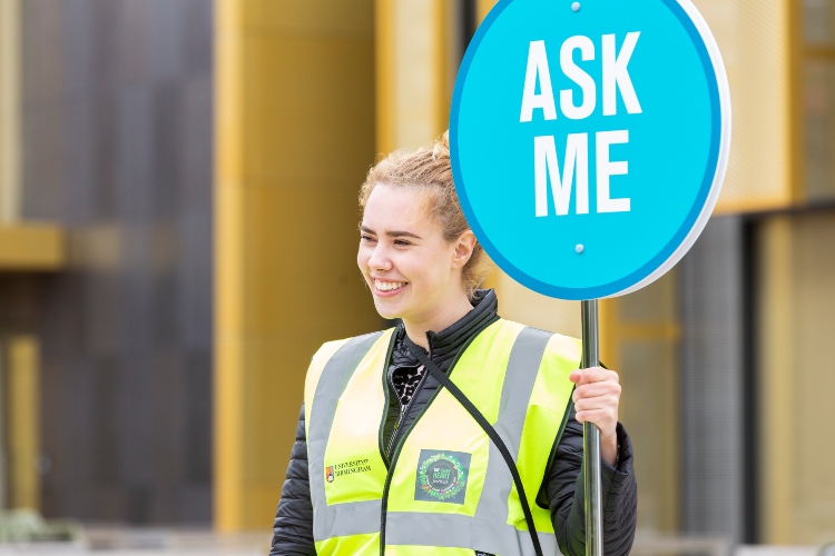 Smiling student holding a blue 'Ask me' sign at a campus event