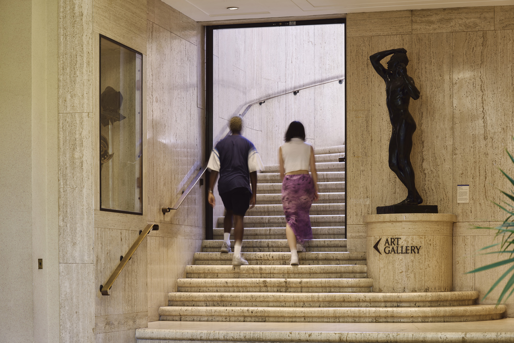 Two people walking up some stairs at the Barber Gallery with a sign pointing to the art gallery 