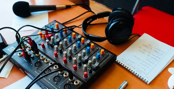 A desk with a small audio mixer which has a microphone and a pair of headphones plugged into it. A pad and pen is also on the desk