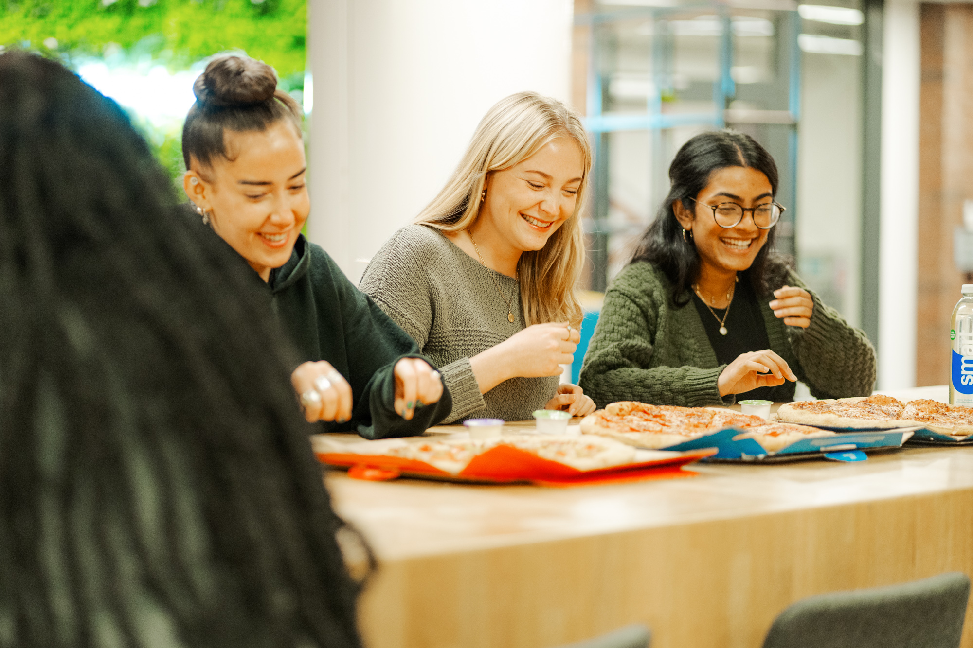 Three students laughing while eating pizza together.