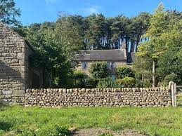 A house in the countryside with a stone wall in front of it and trees surrounding it.