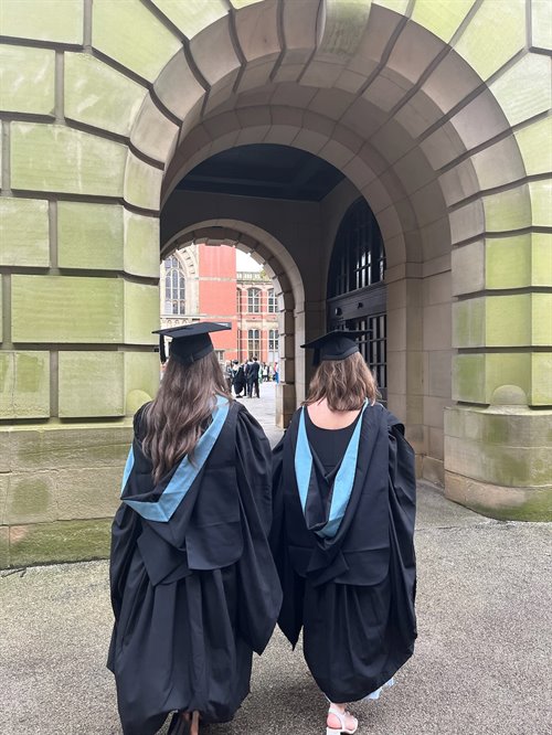 Two female students wearing a graduation cap and gown stand with their back towards the camera