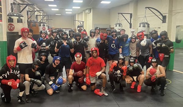 A mix of over 20 members of the UoB Boxing team doing various poses in the gym to the camera. The members are all wearing head gear and boxing gloves.