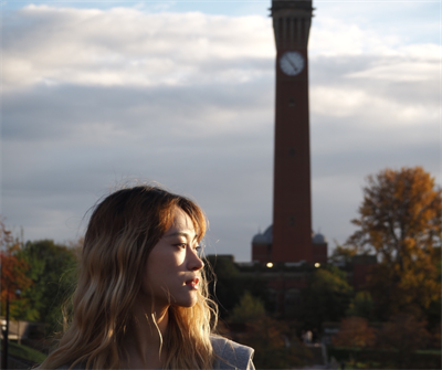 Female student with blonde hair stands with her face turned to the right. Behind her is a tall brick clock tower and trees with golden leaves