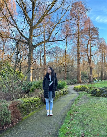 A girl in a coat stands at the top of a path in a green park area.