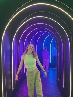 Caitlin is wearing cargo trousers and a vest top. She's smiling at the camera, being photographed in a walkway with neon lighting.