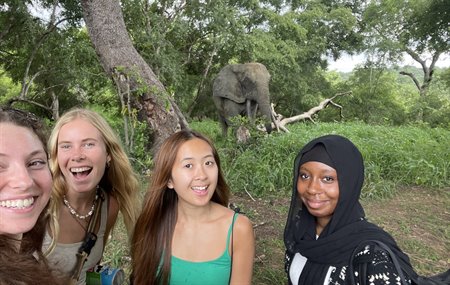 Four girls smile at the camera with an elephant in the background.