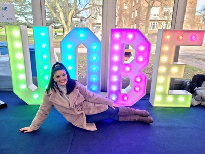 Katrina poses smiling next to a light up sign that reads 'UoBe'.