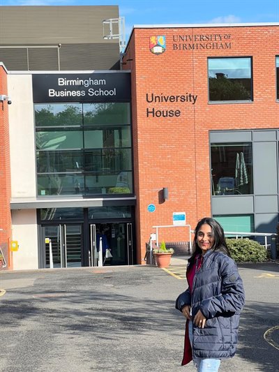 A girl in a blue jacket stands at the front of the photo with the University of Birmingham Business School in the background