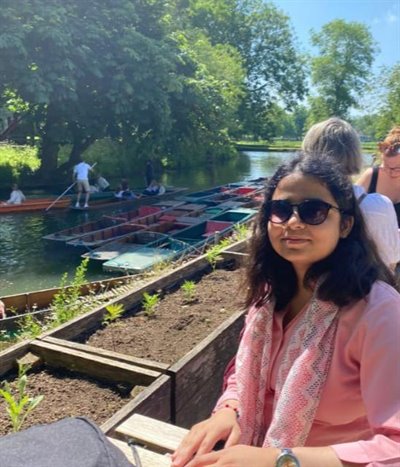 A girl in a pink top and sunglasses smiles at the camera by a river.