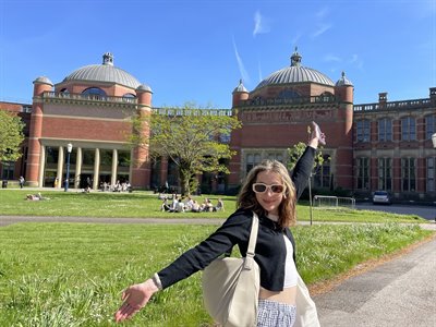 Yael stands outside of the Aston Webb buildings on a sunny day, her arms wide as if to display the buildings behind her,