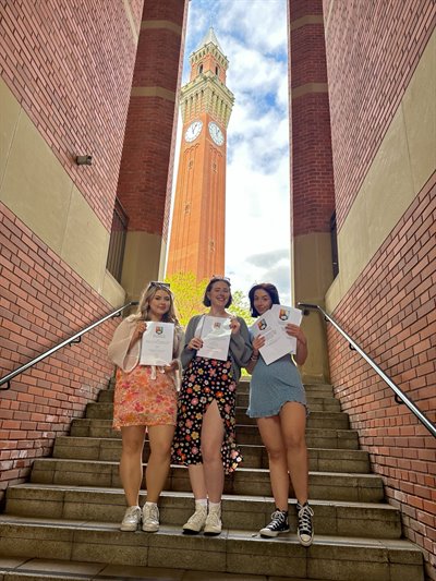 Three girls stand at the bottom of some university steps, holding up their printed dissertations. Old Joe is visible in the background behind them.