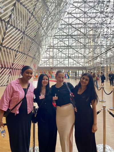 Four girls stand posed together at the front of the photo. The background is a complex, white indoor structure of the building they are in.