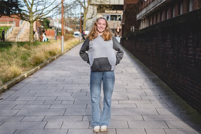 Female student is standing wearing a grey jumper and blue jeans smiling to the camera