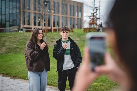 A male and female student, both in black coats, are being filmed for a video in front of the Teaching and Learning building.