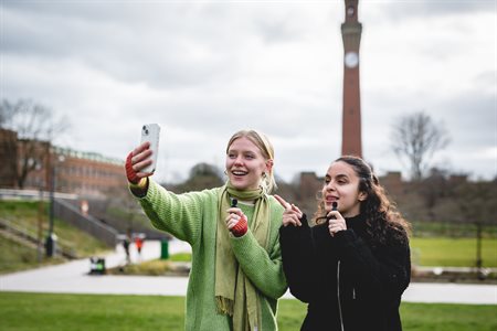 Two girls, one in a green jumper and one in a black coat, film a video in front of Old Joe.