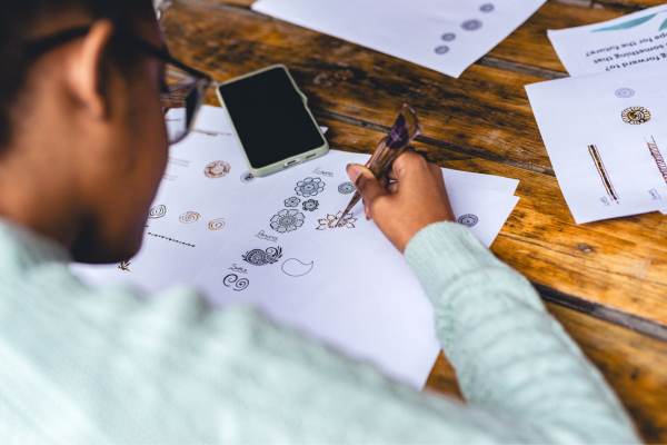 A female student is holding a henna cone and practising a flower design on a white piece of paper