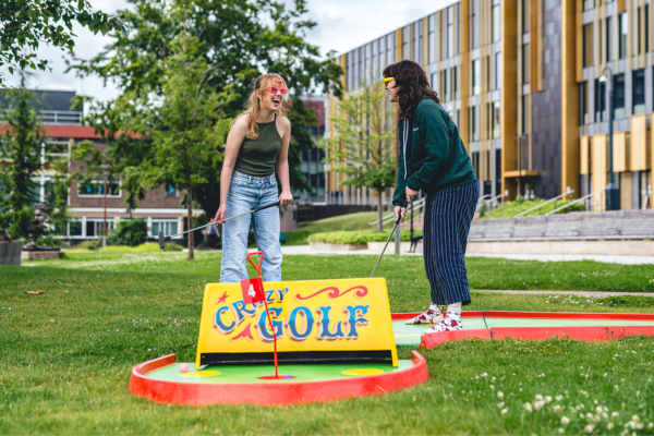 Two female students are facing each other and laughing. They are both holding golf clubs and standing next to a mini golf sign